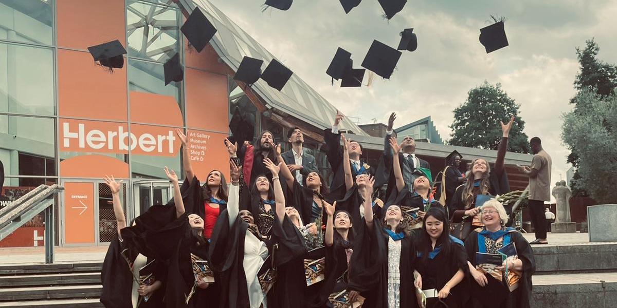 A group of CU Coventry students celebrating their graduation day by throwing their caps in the air.